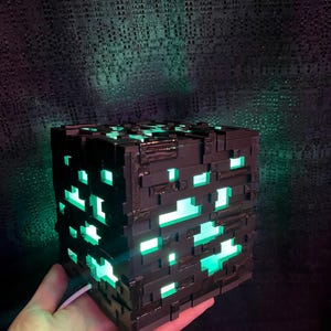 May include: A black, cube-shaped lamp with a pixelated design. The lamp has bright green lights emanating from within, creating a glowing effect. The lamp is held in a hand, with a white cord extending from it.