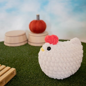 May include: A white crocheted chicken plush toy with a red comb and a yellow beak. The toy is sitting on a green artificial grass surface.