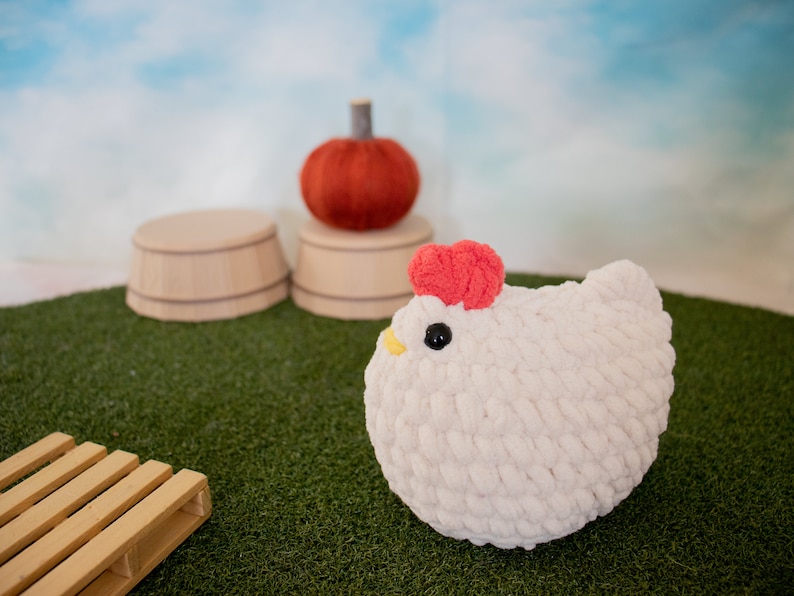 May include: A white crocheted chicken plush toy with a red comb and a yellow beak. The toy is sitting on a green artificial grass surface.