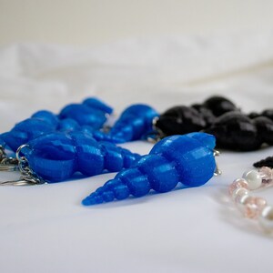Op de afbeelding: Een set blauwe en zwarte schelpvormige sleutelhangers met zilveren sleutelringen. De blauwe schelpen hebben een glinsterende afwerking.