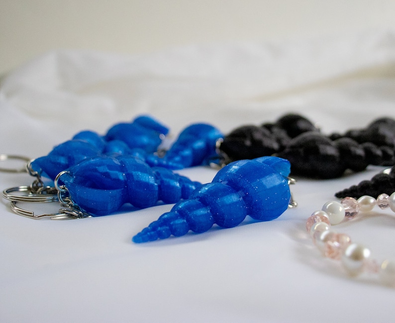 Op de afbeelding: Een set blauwe en zwarte schelpvormige sleutelhangers met zilveren sleutelringen. De blauwe schelpen hebben een glinsterende afwerking.