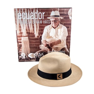 May include: A tan Panama hat with a black band and a brown leather logo. The hat is sitting on a white background. The hat is made in Ecuador and is called a "toquilla straw hat".