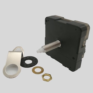 Könnte beinhalten: Ein schwarzes Uhrwerk mit einem silbernen Gewindestab und weißer Spitze, zusammen mit einem Metallbügel, einer Gummischeibe, einer Messingscheibe und einer Messingmutter. Für die Reparatur oder den Bau von Uhren.