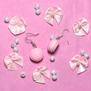 Könnte beinhalten: Ein Paar rosa Makronen-Ohrringe mit silbernen Haken. Die Ohrringe sind von rosa Schleifen und weißen Perlen umgeben.