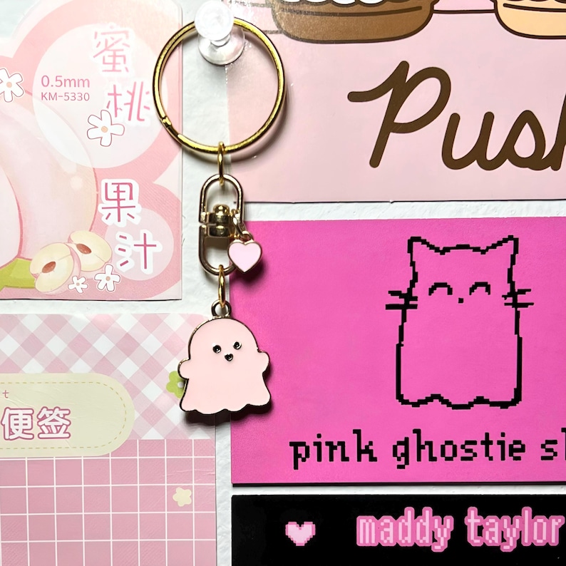 May include: A gold-coloured keyring with a pink ghost charm, a small pink heart charm, and a gold ring. The background features pink and white paper with Japanese text and the words "pink ghostie sl" and "maddy taylor".