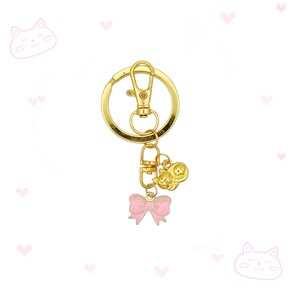 May include: A gold-colored keychain with a lobster clasp and a split ring. The keychain features a small gold cat charm and a pink bow charm. The background is white with pink cat and heart illustrations.