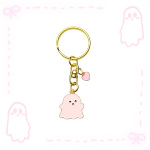 May include: A gold-coloured keyring with a pink ghost charm and a small pink heart charm. The ghost charm has a white outline and small black eyes. The keyring is on a white background with pink ghost and bow illustrations.