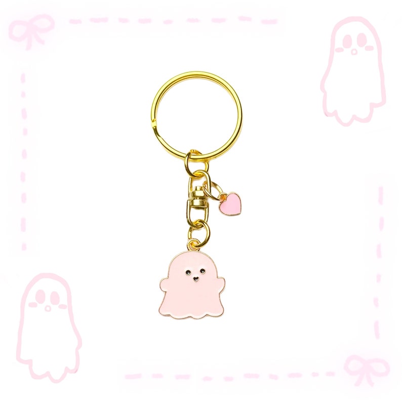 May include: A gold-coloured keyring with a pink ghost charm and a small pink heart charm. The ghost charm has a white outline and small black eyes. The keyring is on a white background with pink ghost and bow illustrations.