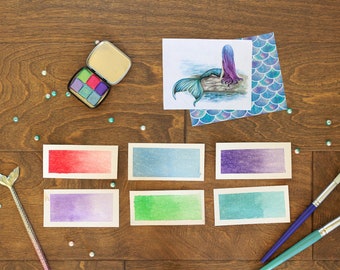 May include: A set of six watercolor paint swatches in shades of red, blue, purple, green, and teal. The swatches are arranged in two rows of three. There is also a mermaid illustration, a paint palette, and two paintbrushes.