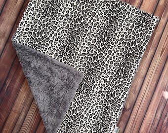 May include: A soft, square blanket with a black and white leopard print pattern. The blanket has a gray, plush backing. The blanket is folded on a wooden surface.