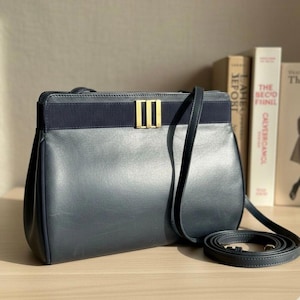May include: A navy blue leather handbag with a gold-colored rectangular clasp. The bag has a shoulder strap and a dark blue fabric band across the top. The bag is sitting on a wooden surface.