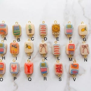 May include: A collection of rectangular charms with gold-colored loops. The charms feature various designs, including a sun, a lemon, a cactus, and a lobster. The charms are connected to colorful cords, suggesting they are for jewelry.