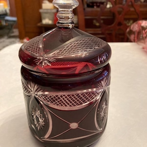 May include: A ruby red, cut glass jar with a lid. The jar features intricate starburst and geometric patterns. The lid has a clear, rounded knob. The jar is likely used for storing small items or as a decorative piece.