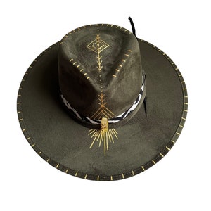 Puede incluir: Un sombrero vaquero de gamuza verde con costuras doradas y un diseño geométrico en la corona. El sombrero tiene una pluma negra y una banda con estampado blanco y negro.