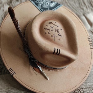 May include: A tan felt fedora hat with a black and white paisley bandana band. The hat has a feather attached to the side and a variety of symbols drawn on the crown.