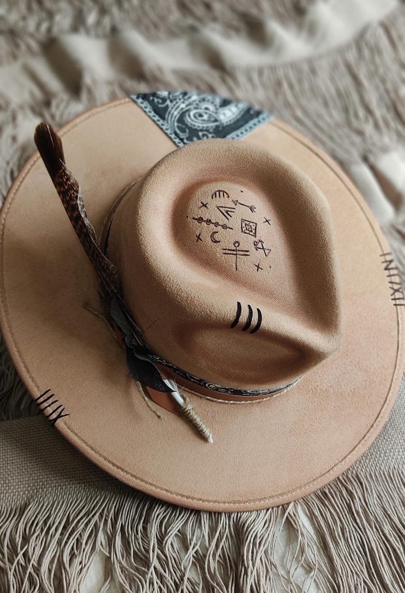 May include: A tan felt fedora hat with a black and white paisley bandana band. The hat has a feather attached to the side and a variety of symbols drawn on the crown.