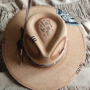 May include: A brown suede cowboy hat with a black bandana and a feather on the side. The hat has a stitched design around the brim and a symbol drawn on the crown.