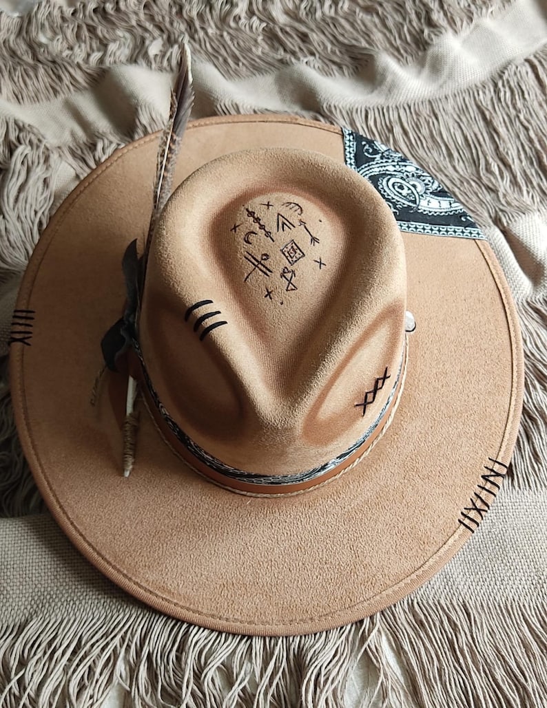 May include: A brown suede cowboy hat with a black bandana and a feather on the side. The hat has a stitched design around the brim and a symbol drawn on the crown.
