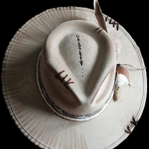 May include: A beige cowboy hat with a brown leather band and a feather accent. The hat has a stitched design on the crown and a brown leather band with a white and black patterned design.