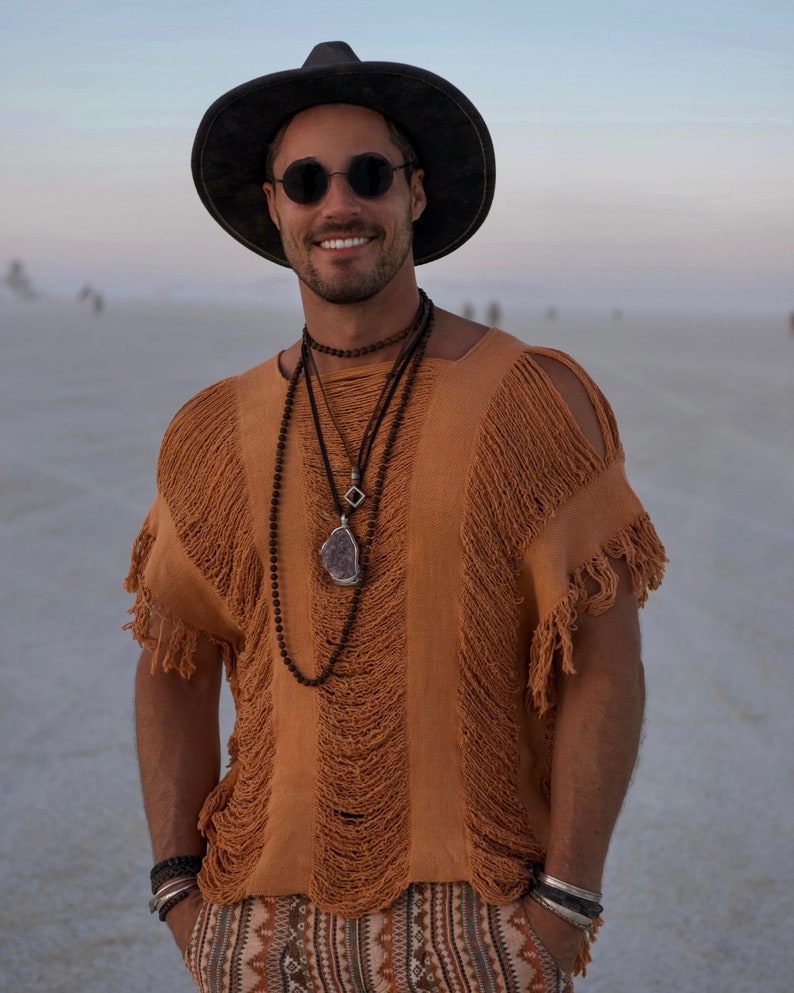 Puede incluir: Un hombre con un sombrero negro de ala ancha, gafas de sol y una camisa marr&oacute;n con flecos y varios collares. Est&aacute; de pie en un paisaje des&eacute;rtico.