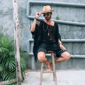 Puede incluir: Un hombre con un sombrero marr&oacute;n con una pluma, gafas de sol y un poncho negro con flecos se sienta en un taburete de madera. Lleva pantalones cortos negros y un collar de cadena de plata con un colgante de cristal.