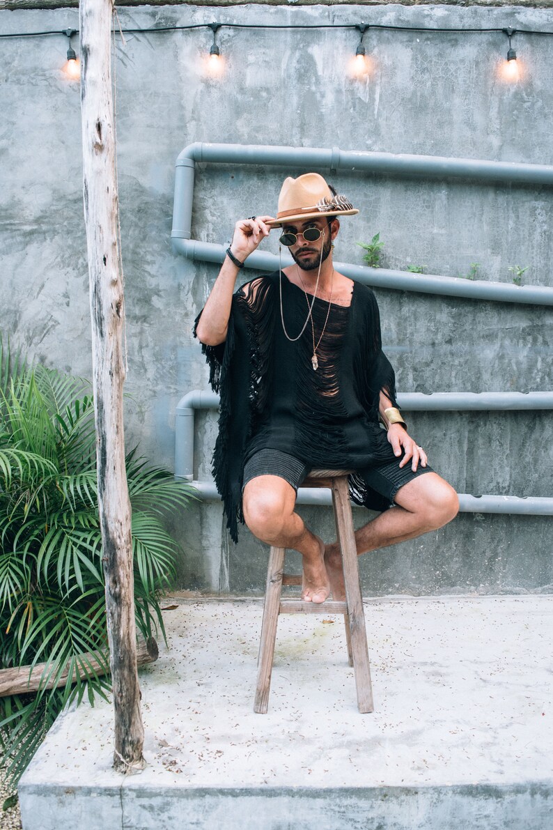 Puede incluir: Un hombre con un sombrero marr&oacute;n con una pluma, gafas de sol y un poncho negro con flecos se sienta en un taburete de madera. Lleva pantalones cortos negros y un collar de cadena de plata con un colgante de cristal.