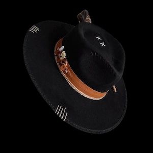 Könnte beinhalten: Schwarzer Fedora-Hut mit breiter Krempe. Er hat ein braunes Lederband mit dekorativen Nähten und kleinen Verzierungen. Weiße Kreuzstichdetails befinden sich auf der Krone. Eine Feder ist an der Seite befestigt.