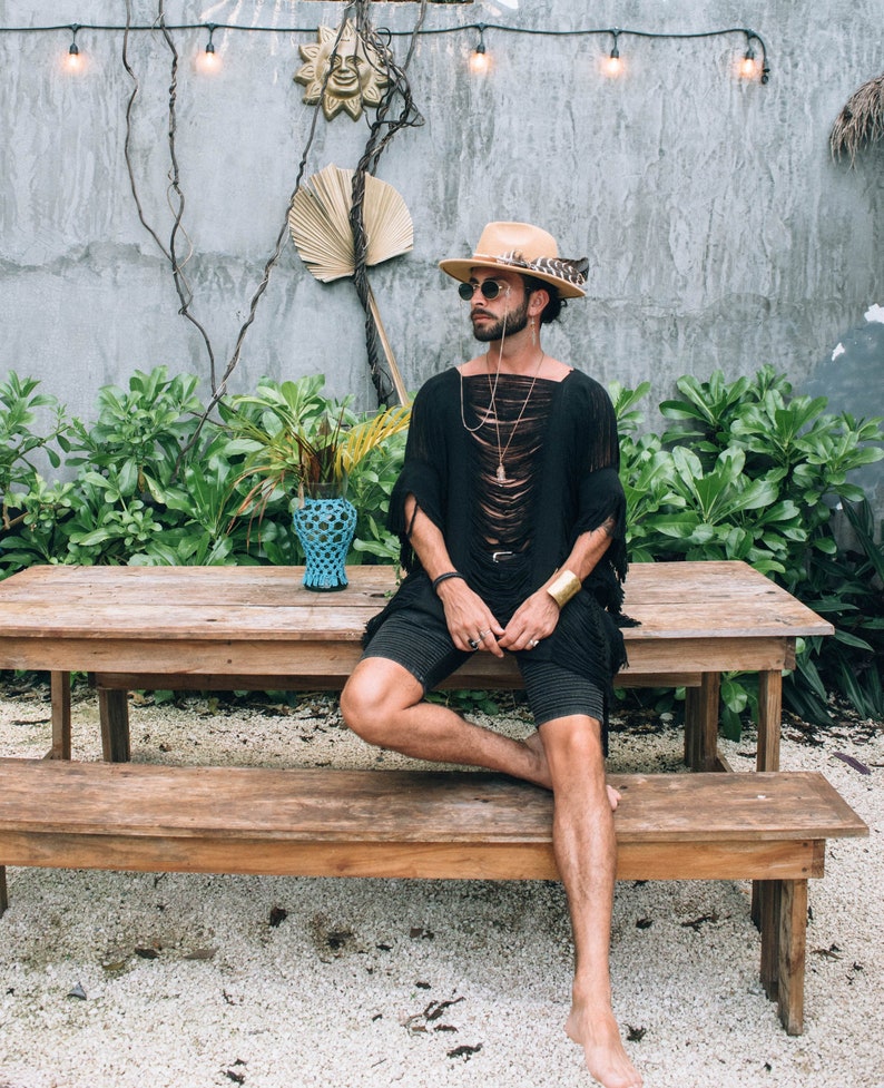 Puede incluir: Una persona con una camisa negra de manga corta desgarrada, pantalones cortos negros, un sombrero marr&oacute;n con una pluma y gafas de sol se sienta en un banco de madera. El banco est&aacute; delante de una mesa de madera. El fondo es una pared gris con luces de cadena y vegetaci&oacute;n.