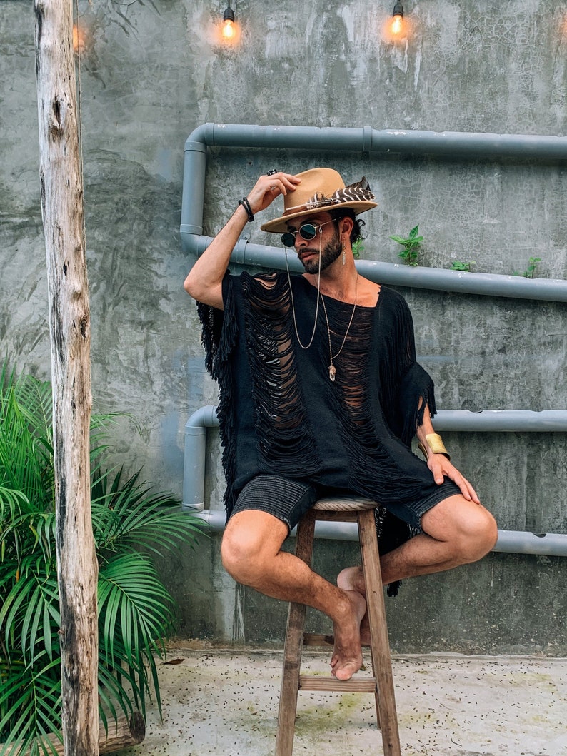 Puede incluir: Un hombre con una camisa negra de manga corta desgarrada, pantalones cortos marrones, un sombrero marr&oacute;n con una pluma y gafas de sol est&aacute; sentado en un taburete de madera.