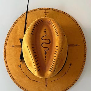 Könnte beinhalten: Senffarbener Cowboyhut mit schwarzer Feder und dekorativem, genähtem Rand. Der Hut hat ein schwarzes Schlangenmuster auf der Krone und Pfeil- und Kreuzmuster auf dem Rand. Ein stilvolles Accessoire.