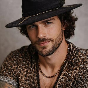May include: A black fedora hat featuring gold-colored geometric embroidery along the crown and brim, accented with a dark feather. The hat is worn with a brown and black patterned short-sleeve shirt and a layered beaded necklace with a small pendant.