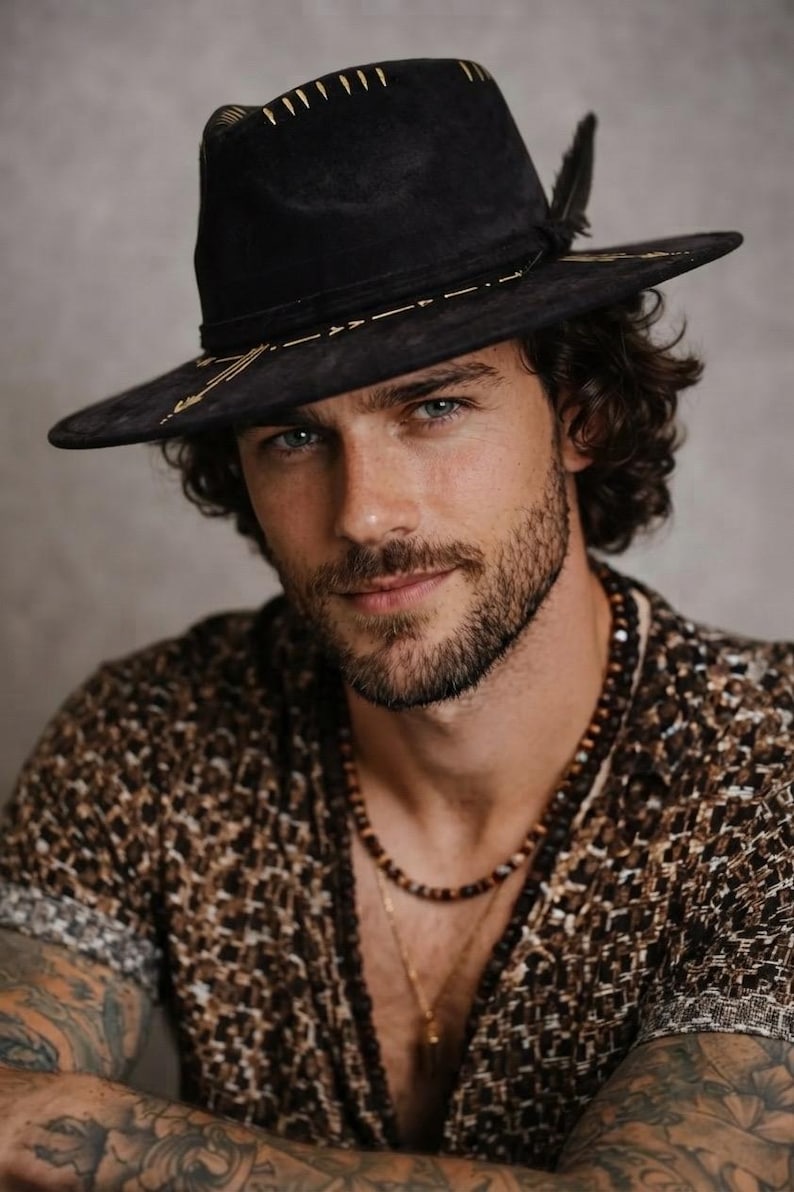 May include: A black fedora hat featuring gold-colored geometric embroidery along the crown and brim, accented with a dark feather. The hat is worn with a brown and black patterned short-sleeve shirt and a layered beaded necklace with a small pendant.