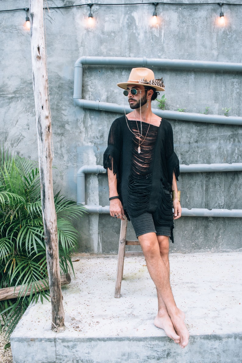 Puede incluir: Una persona con un top negro deshilachado con flecos, un sombrero marr&oacute;n, gafas de sol y varios collares. La persona est&aacute; de pie en una plataforma de hormig&oacute;n con un taburete de madera.