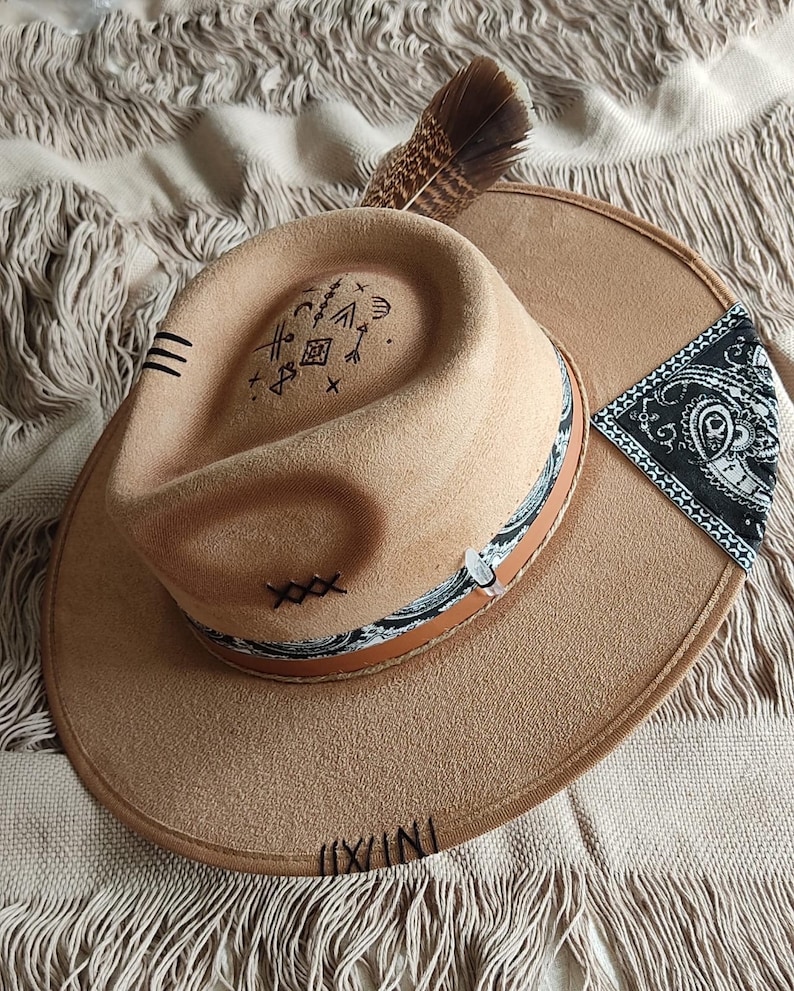 May include: A brown felt fedora hat with a black and white patterned band, a feather accent, and various symbols embroidered on the crown. The hat is sitting on a white fringed fabric.