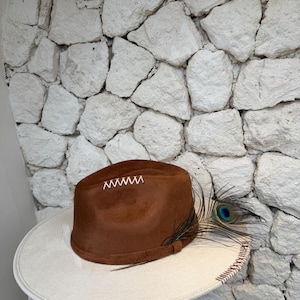 Puede incluir: Un sombrero fedora marrón con un ala ancha de color crema que presenta costuras de color marrón oscuro a lo largo del borde. La copa está adornada con bordados blancos en zigzag y una pluma de pavo real insertada en una cinta marrón.