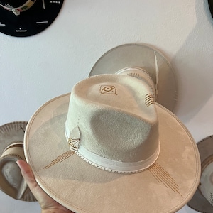 May include: A light beige fedora-style hat with a wide brim. The hat features a white band with small silver studs, gold-colored decorative lines, and a small white stone accent. The hat is displayed against a white wall with other hats.