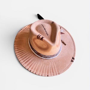 Könnte beinhalten: Ein beigefarbener Fedora-Hut mit breiter, plissierter Krempe. Der Hut hat dekorative Nähte auf der Krone und ein gemustertes Band um den Ansatz. Ein schwarzer Akzent ist am Hut befestigt.