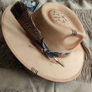 May include: A tan felt fedora hat with a black and white bandana tied around the crown. A brown feather is attached to the hat with a black ribbon. The hat has a black design on the top that includes symbols and lines.