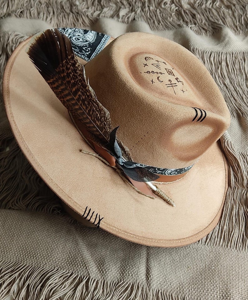 May include: A tan felt fedora hat with a black and white bandana tied around the crown. A brown feather is attached to the hat with a black ribbon. The hat has a black design on the top that includes symbols and lines.