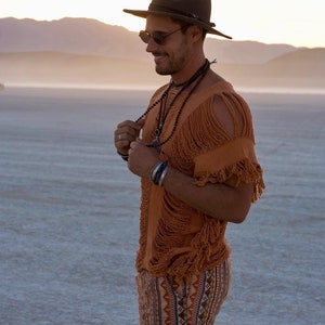 Puede incluir: Un hombre con una camisa marr&oacute;n con flecos, pantalones cortos negros con un patr&oacute;n geom&eacute;trico, un sombrero negro y gafas de sol. Est&aacute; de pie en un paisaje des&eacute;rtico.