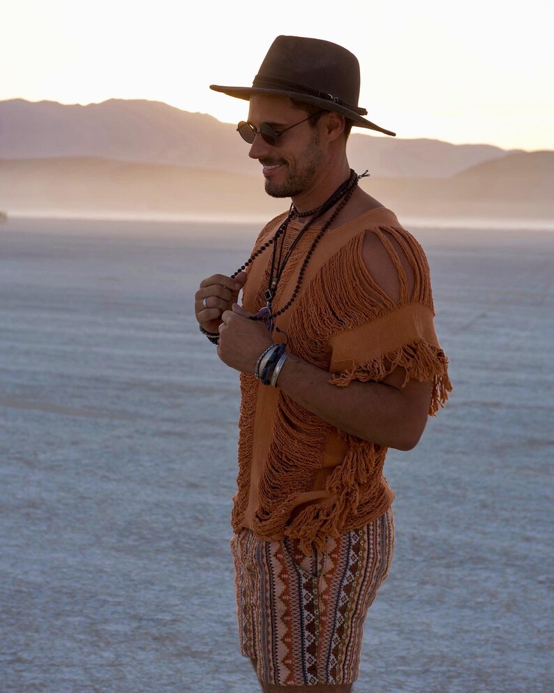 Puede incluir: Un hombre con una camisa marr&oacute;n con flecos, pantalones cortos negros con un patr&oacute;n geom&eacute;trico, un sombrero negro y gafas de sol. Est&aacute; de pie en un paisaje des&eacute;rtico.