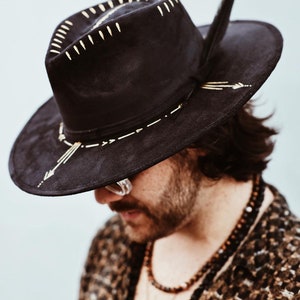 May include: A black wide-brimmed hat featuring gold-coloured painted details on the crown and brim. The hat includes a dark feather tucked into the side band and displays linear, arrow-like, and dash-shaped gold markings across its surface.