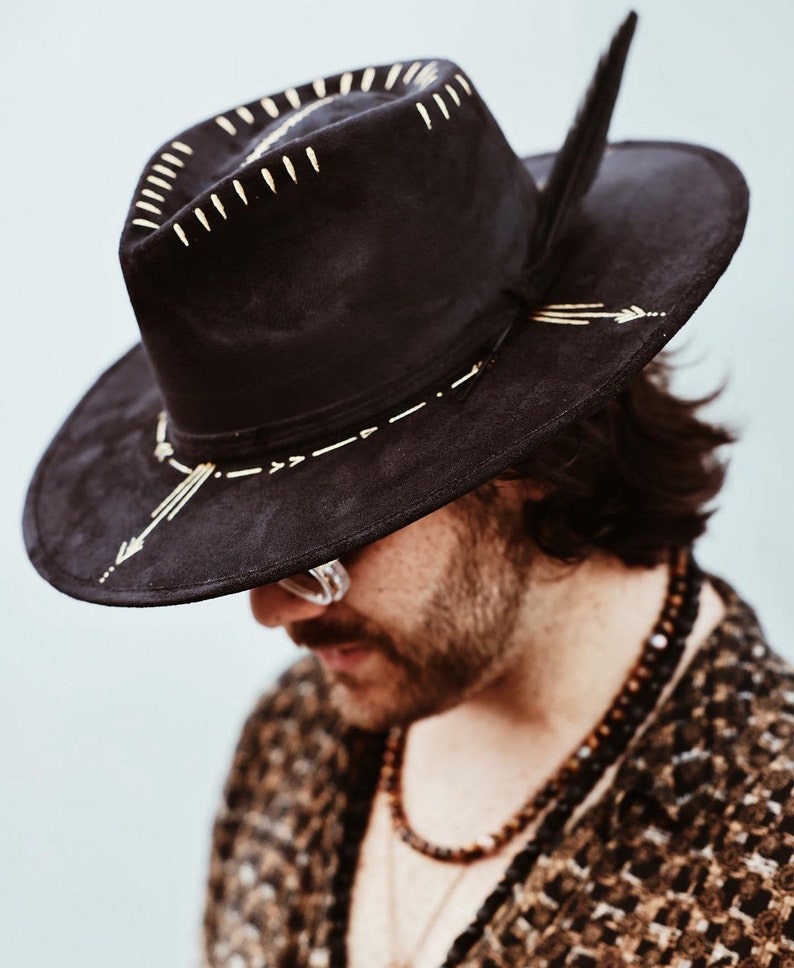 May include: A black wide-brimmed hat featuring gold-colored painted details on the crown and brim. The hat includes a dark feather tucked into the side band and displays linear, arrow-like, and dash-shaped gold markings across its surface.