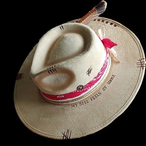 May include: A beige fedora-style hat with a wide brim. The hat features a red and white patterned band, decorative stitching, and a feather accent. The text "MY SOUL FEELS AT HOME" is printed on the brim.