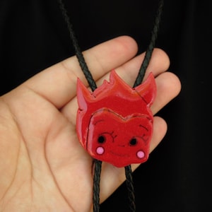 May include: A red devil-faced bolo tie pendant with a smiling expression and pink cheeks. The pendant is attached to a black braided cord. The bolo tie is held in a hand against a black background.