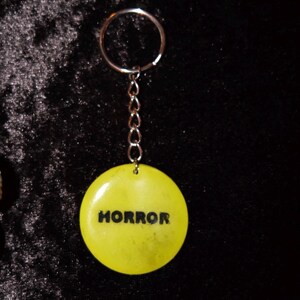 May include: A round, yellow keychain with the word "HORROR" in black capital letters. The keychain has a silver chain and a silver key ring. The background is a dark, textured fabric.