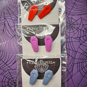 Könnte beinhalten: Drei Paar Sarg-Ohrringe in Rot, Lila und Blau, jeweils mit einem Kreuzdesign. Die Ohrringe sind auf Karten mit dem Text "Bubble Ghoul" und einem dekorativen Logo verpackt. Der Hintergrund zeigt ein lila Spinnennetz-Muster.