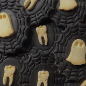 May include: Close-up of Halloween-themed cookies. The cookies are decorated with a black spiderweb design and feature pale yellow ghost and tooth shapes. The cookies are arranged in a circular pattern.
