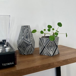 Peut inclure: Deux vases en céramique géométriques noirs et blancs avec un motif géométrique blanc. Le vase de droite contient une petite plante verte. Les vases sont sur une étagère en bois avec un tourne-disque noir à gauche.