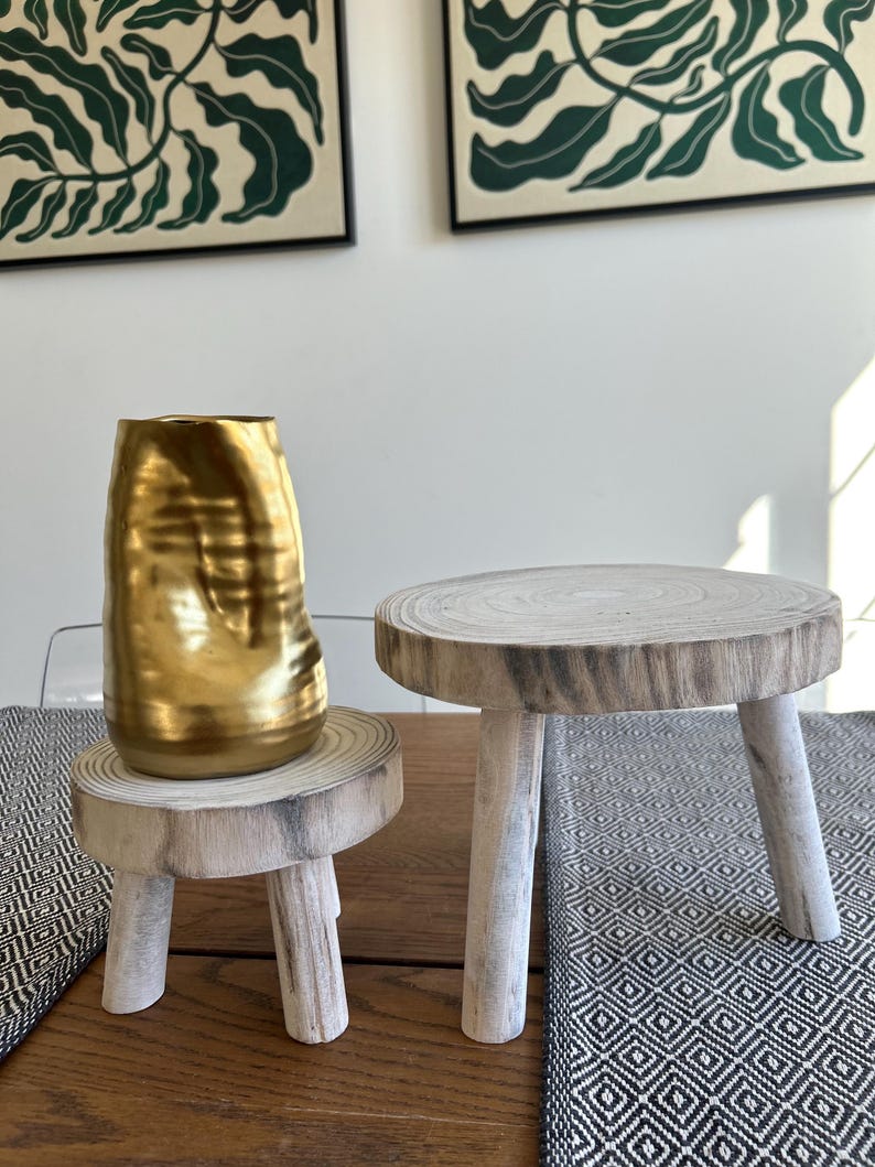 May include: A gold-colored, textured vase sits on a small, round wooden stool with three legs. Beside it is another similar stool. The stools are made of natural wood with a light finish. The background includes framed botanical art.
